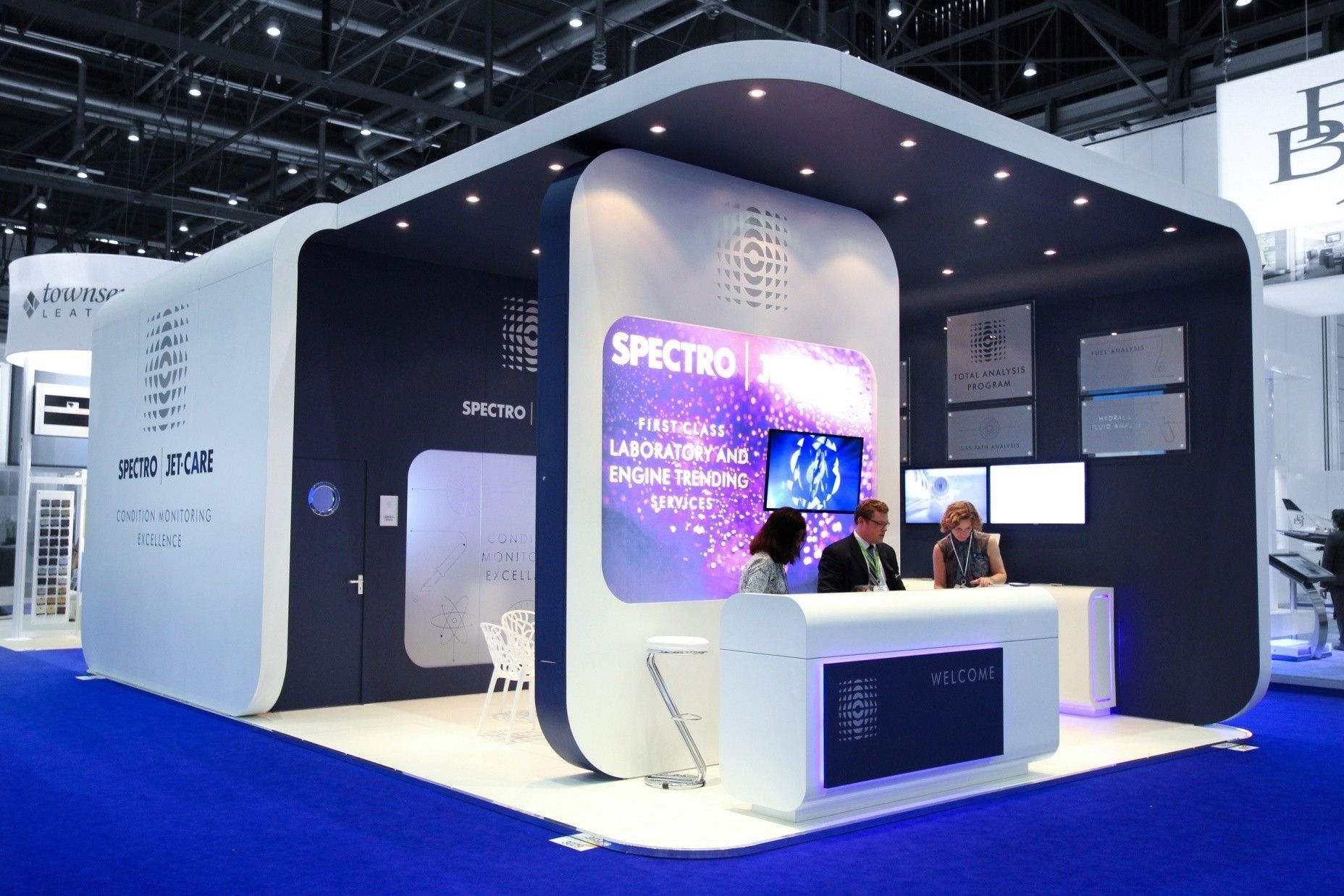 Spectro exhibit booth with two people behind a counter, blue and white colors, bright lights, display screens.
