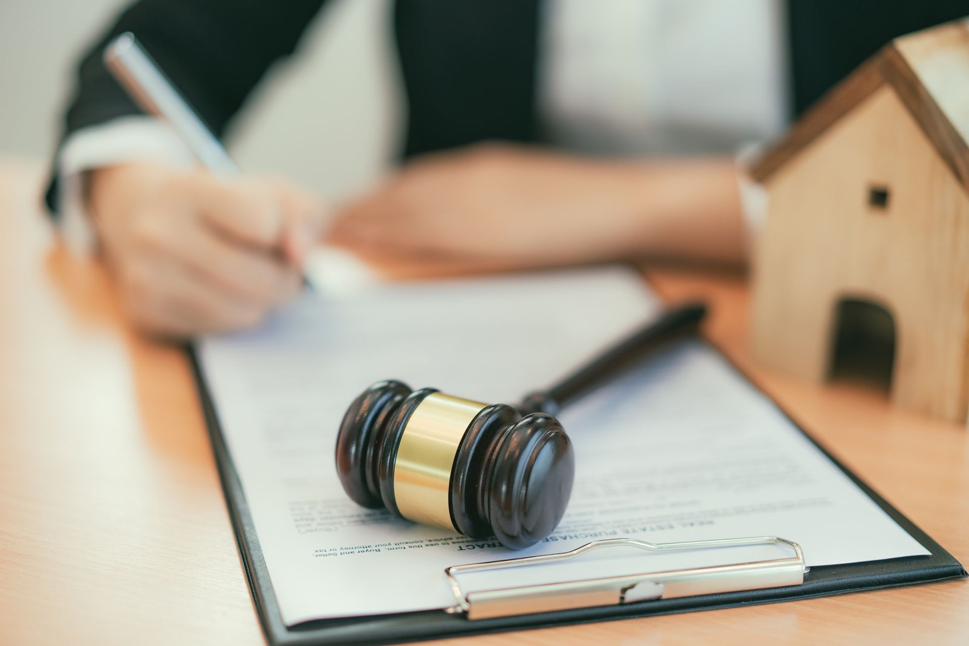 Gavel on clipboard with person writing and house model in background.