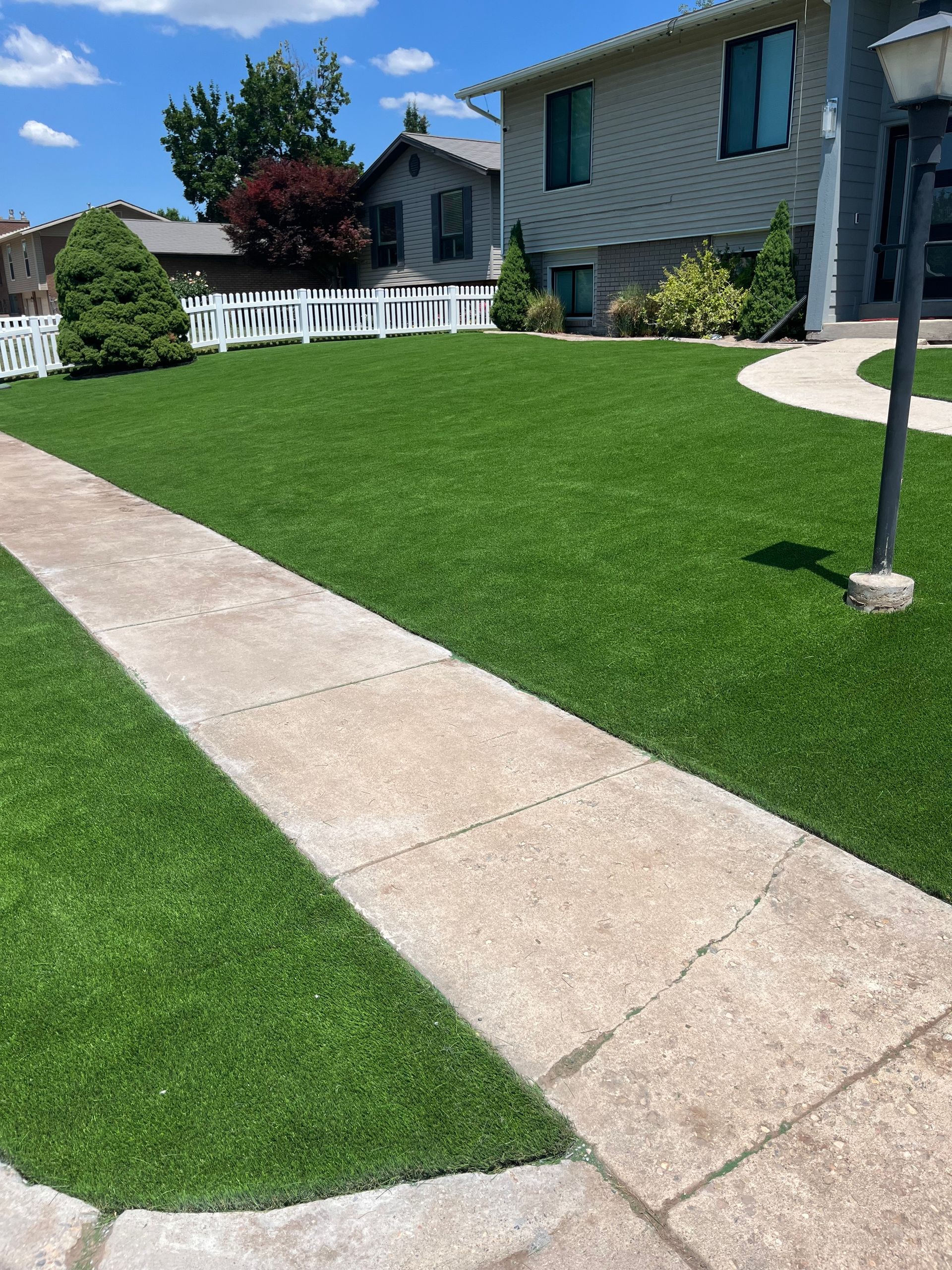 Concrete sidewalk leads to a house with a lush green lawn and a white picket fence. Sunny day.