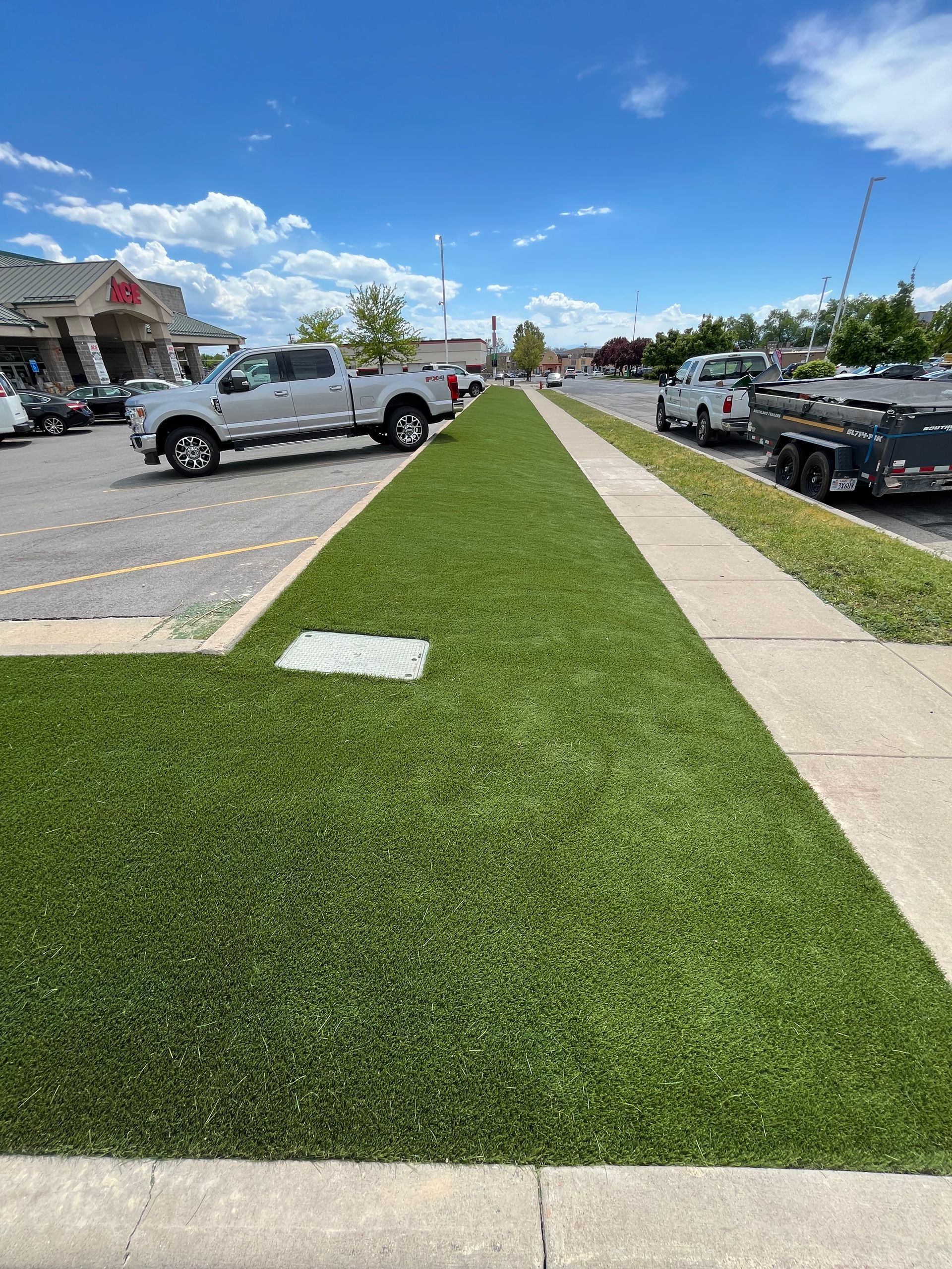 Green artificial turf median strip next to a sidewalk and parking lot with cars.