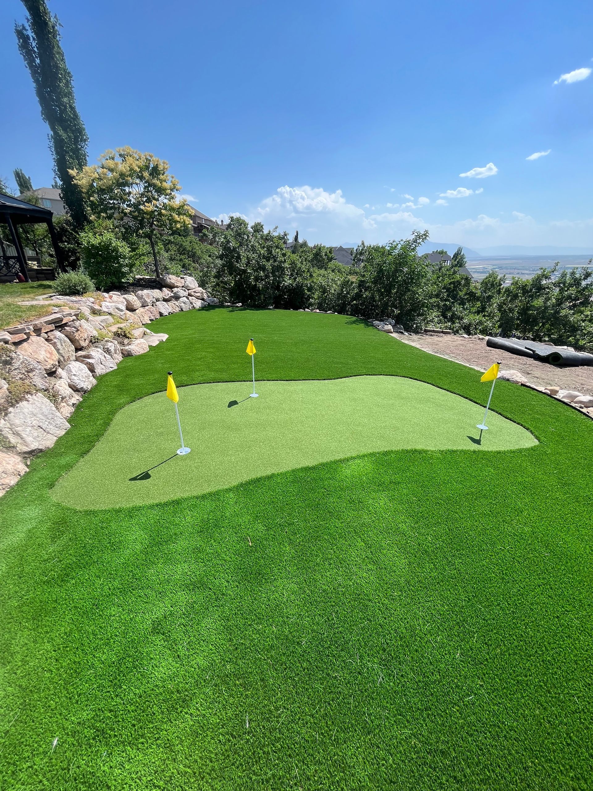 A home putting green with three holes, surrounded by lush artificial grass, under a clear sky.
