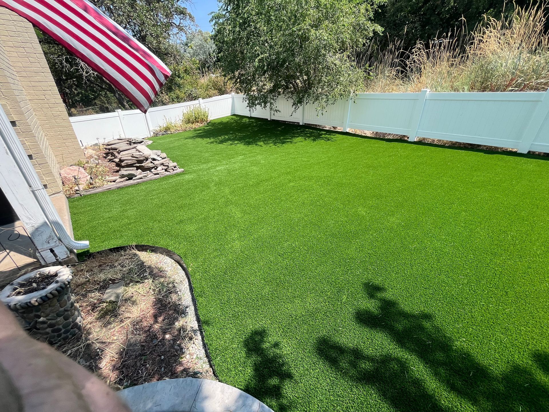 Green artificial lawn in a backyard with white fence, American flag, and small rock garden.