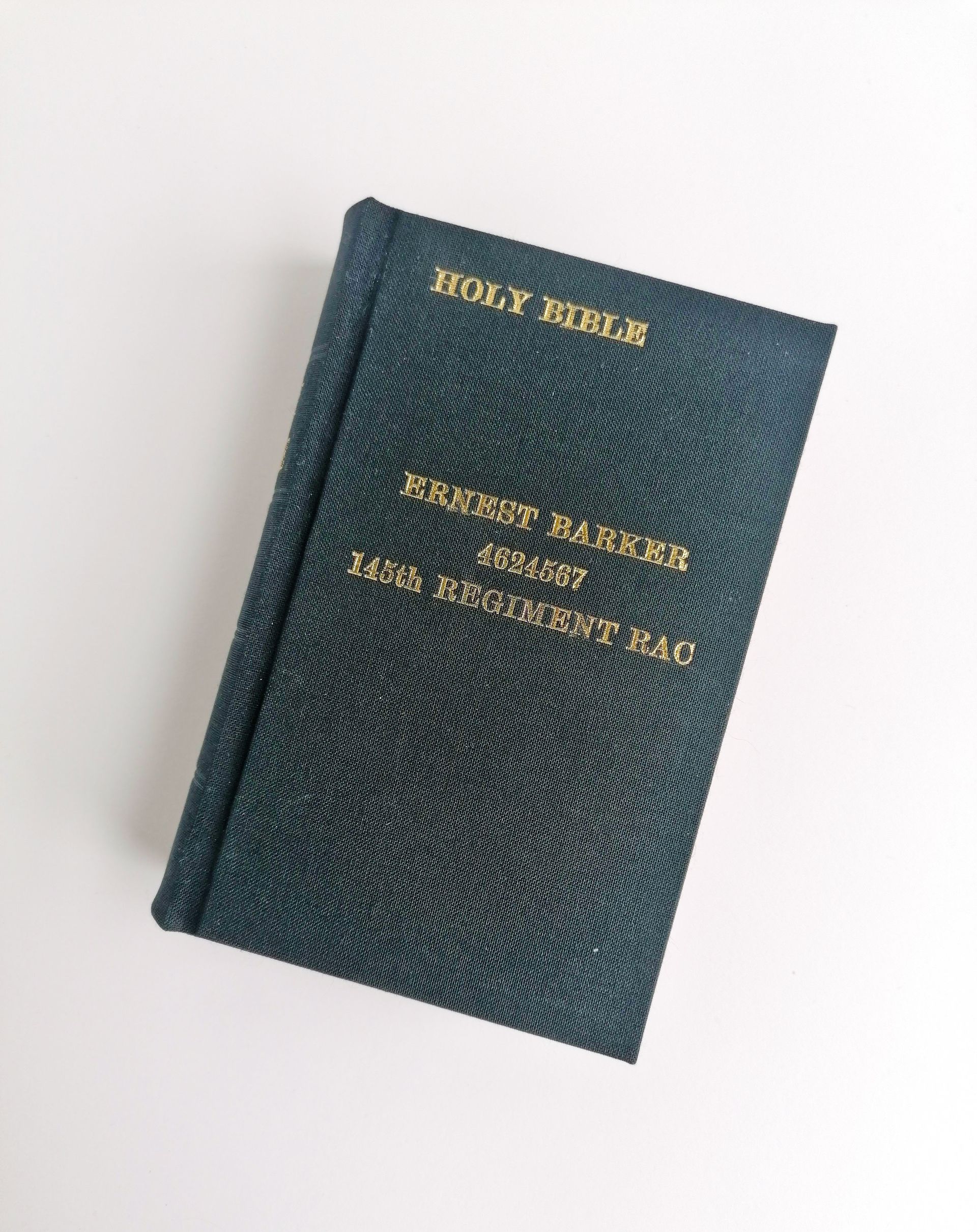 The image shows the fully repaired Holy Bible book. The book now has a new black fabric cover and there are 4 lines of gold text in capitals. The top line reads Holy Bible, and the second third and forth line of text has the owners name, regiment number and the title 145th Regiment RAC. The book sits on a white background.
