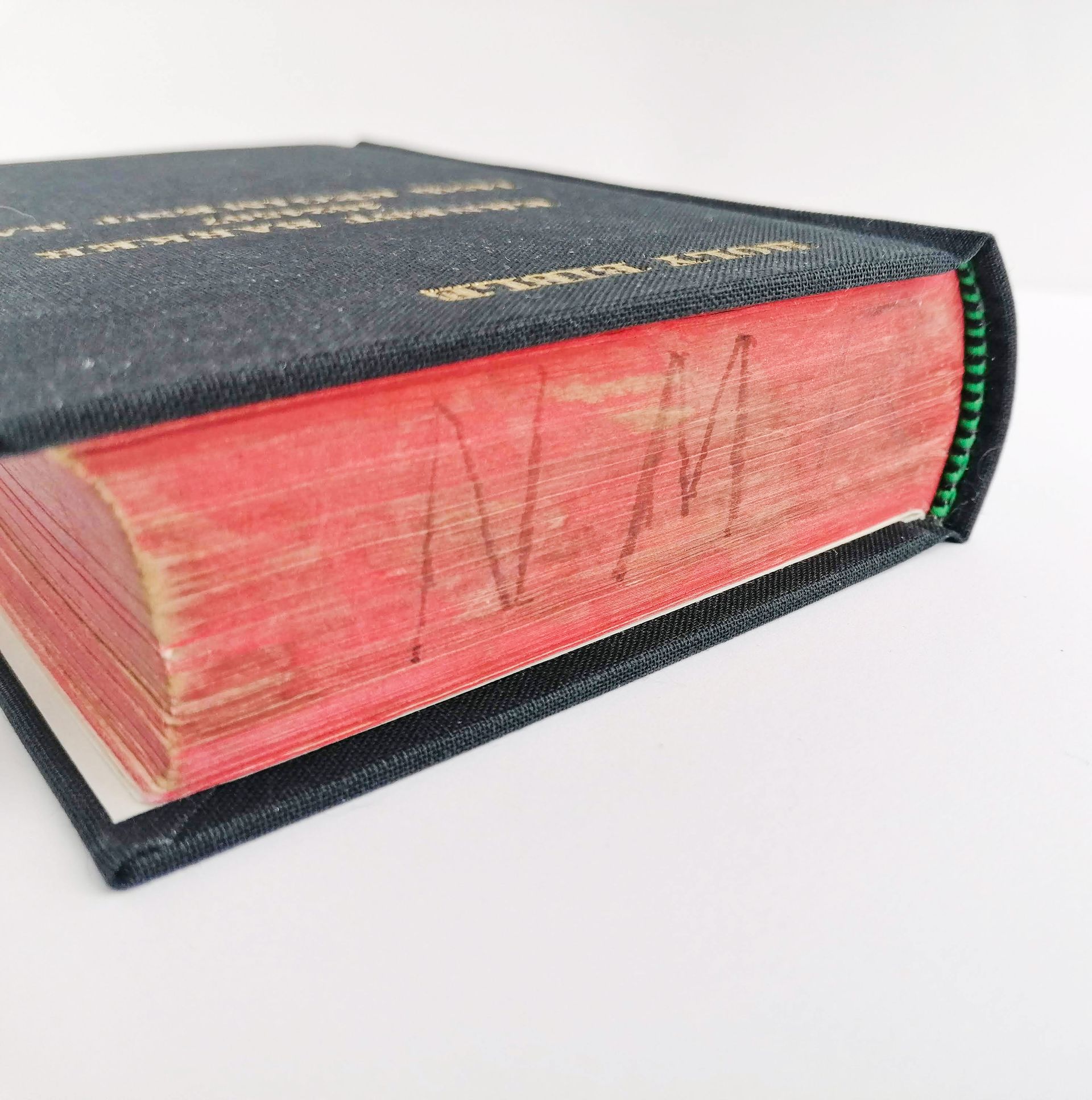 The image shows close up detail of the top of the book. The edges of the pages are red with the initials N. M drawn in pen in capitals, probably from the original owner of the book. The spine is rounded and curved for ease of reading the pages when the book is opened. The book now has a new black fabric cover and the pages have decorative end bands at the top and bottom of the book which are both black and bright green woven colours. The book sits on a white background.