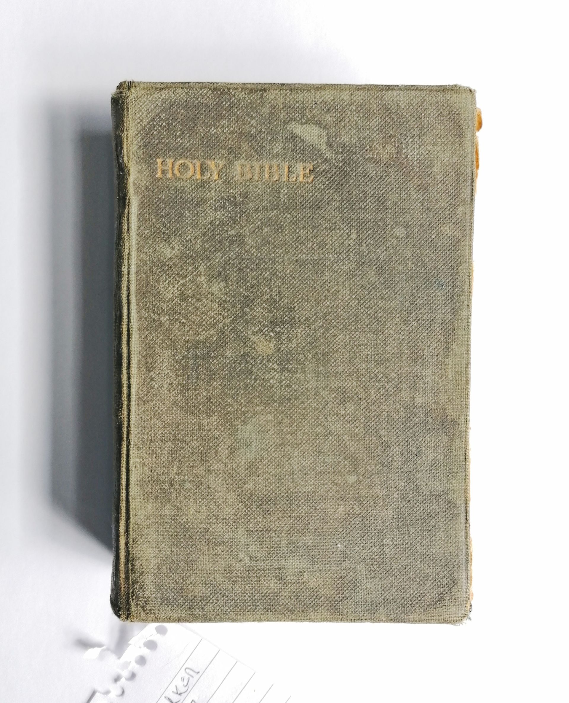 The image shows a an old book on a white background. The front book cover is green fabric on board but the fabric is patchy and faded. The book cover reads Holy Bible in capital gold lettering.