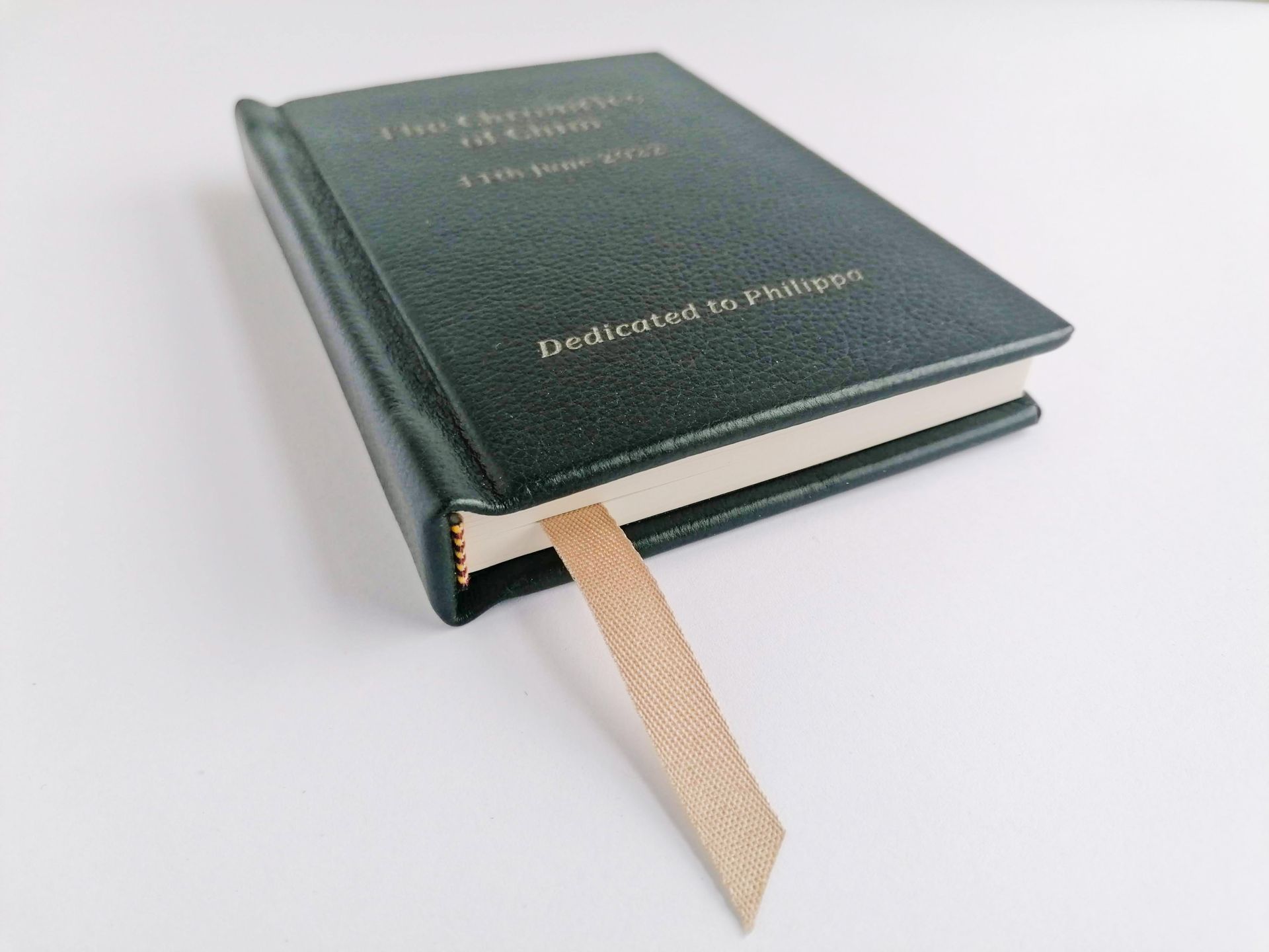 The image shows a small dark green leather book with the cover engraved with a personalised message that reads dedicated to Phillipa. The book has a light tan golden coloured page ribbon and yellow and red decorative traditional end bands at the top and tail of the book.