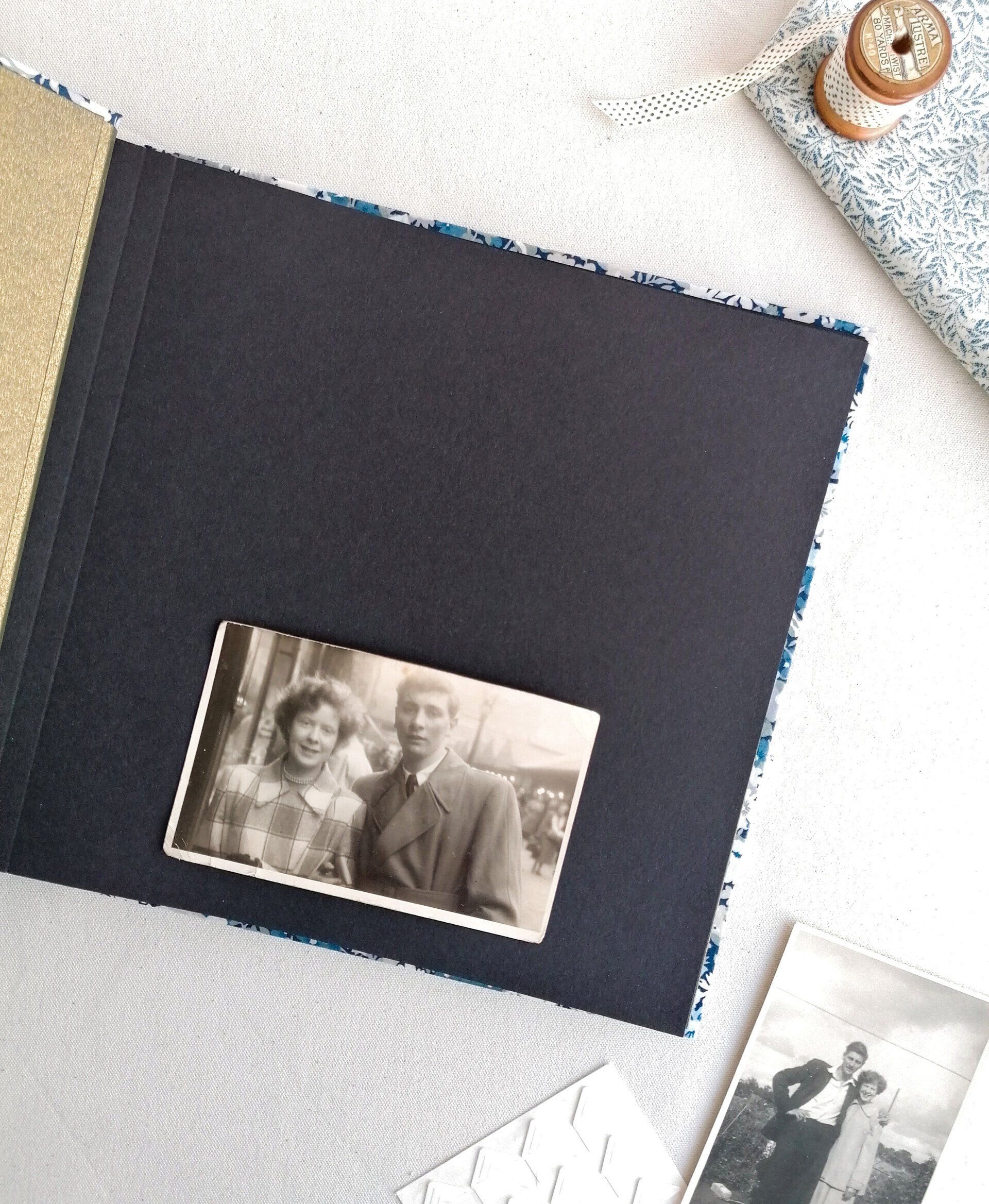 The image shows a floral patterned large landscape photo album which is opened at the first page. The inside of the album cover is gold. The album has black pages and the first page has a vintage photo of a man and woman looking very smart in coats from the 1940's. The album is laid on a white background next to a wooden spool with green spotty ribbon and floral blue and white fabric.