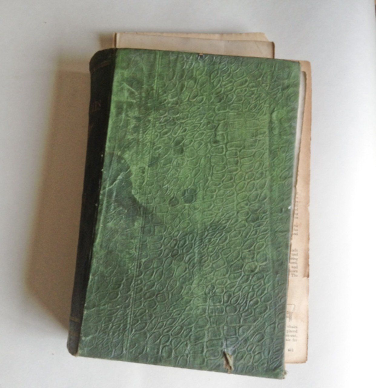 The image shows a damaged book with lots of loose pages falling out. The book covers are dark green and have been water damaged stained and the covering fabric is torn.