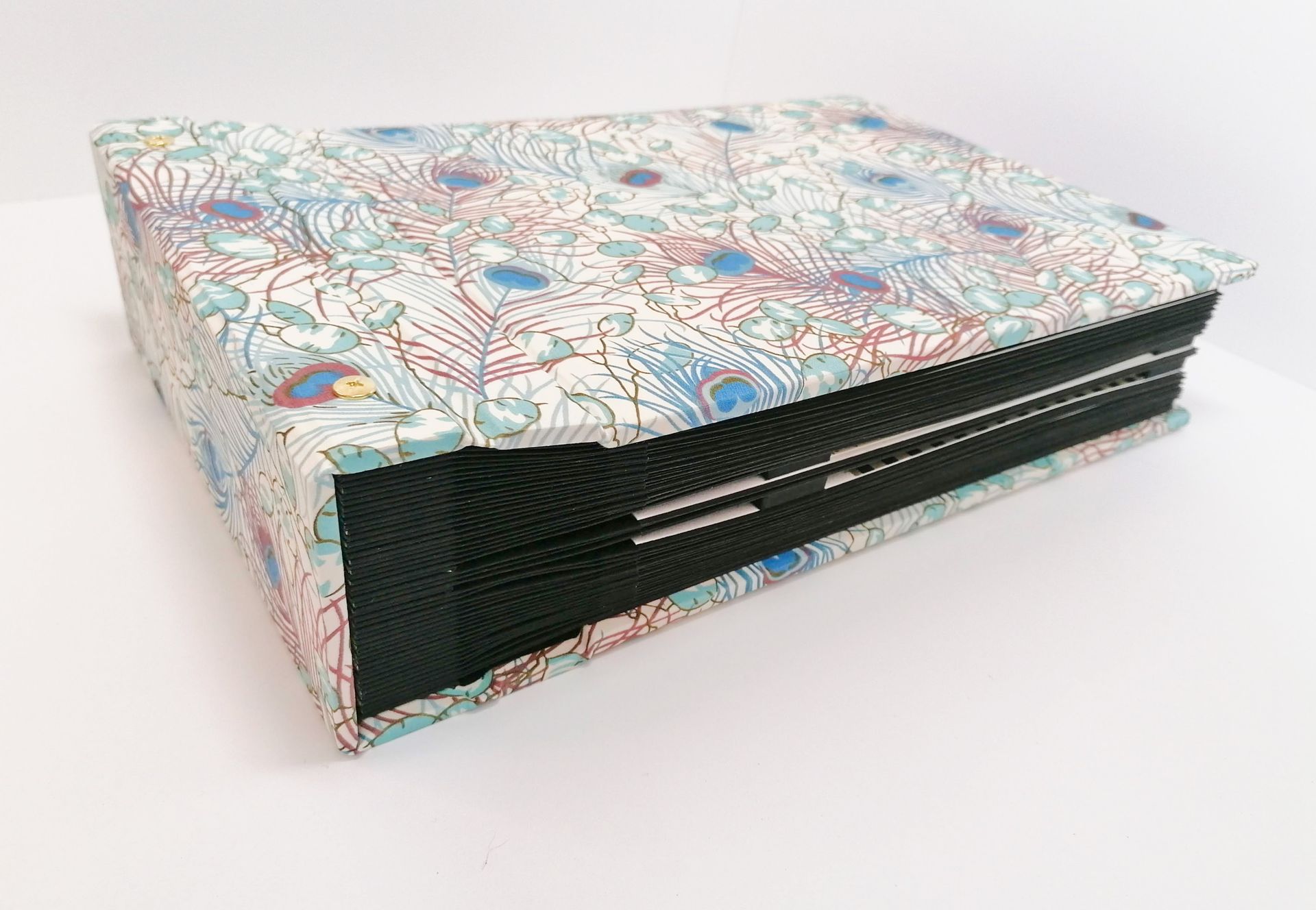 The image shows a very large album from the side with many black coloured pages bound together. The album is covered in a Liberty of London branded fabric which has peacock feathers in turquoise, burgundy and light blue on a white background.