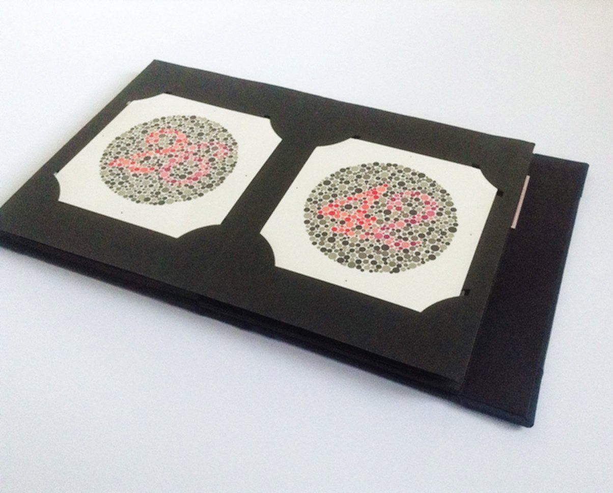 The image shows the dark blue book repaired and laid open flat on the table. The black pages have optical circular patterns on them in green with different coloured circles in red inside the pattern to create numbers. The book is used to test colour blindness.