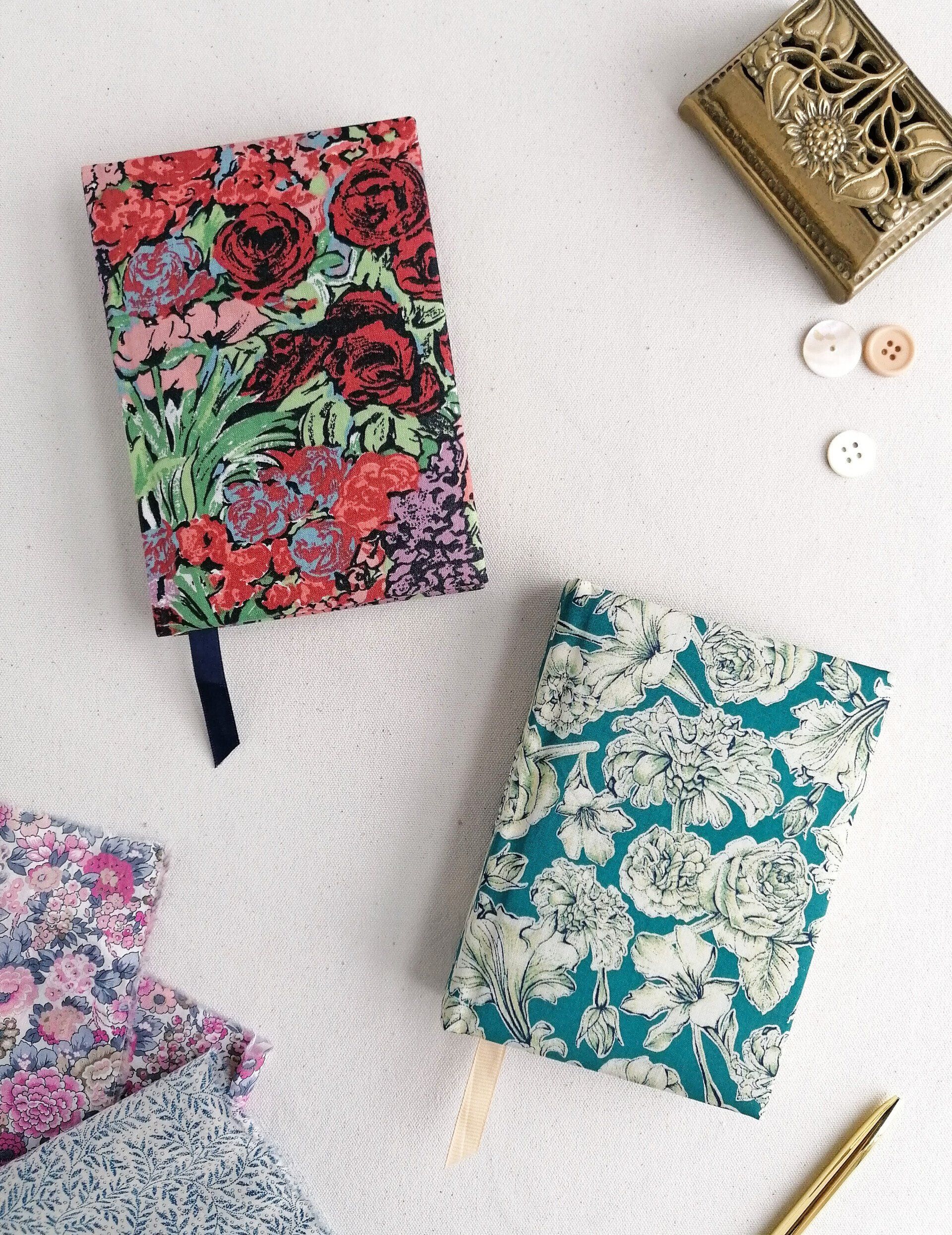 An image of vintage upcycled fabric covered diaries. They are A6 pocket sized and laid on a plain white background. There are 2 diaries with different decorative covers. One is covered in a floral fabric with red pink light purple green and blue flowers. The other diary is covered in a floral fabric with jade light green and white flowers. Both Liberty of London cotton fabrics.