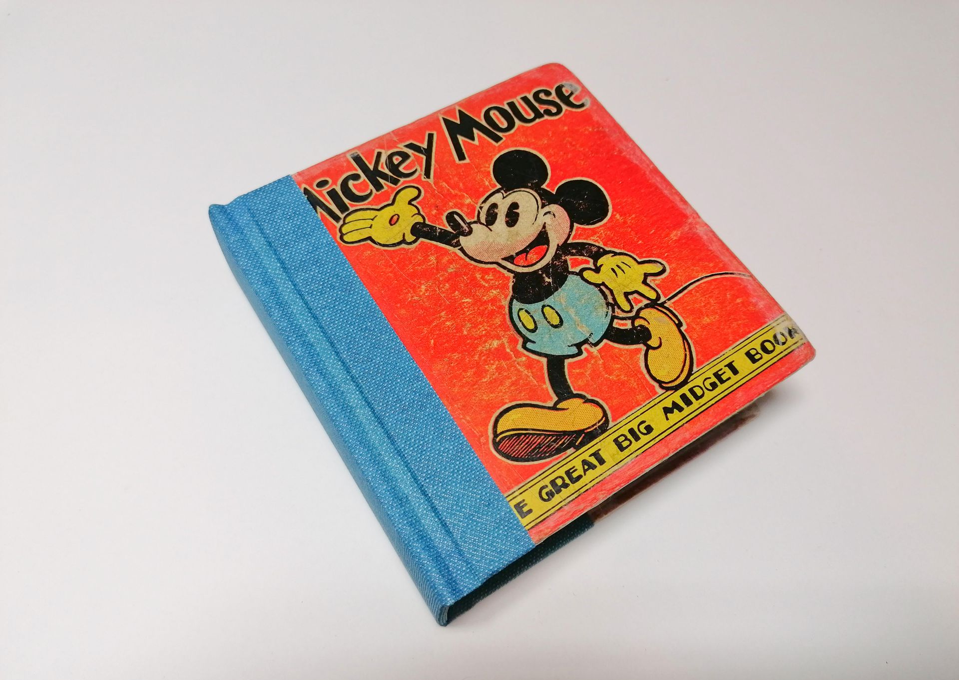 The image shows a repaired small red vintage book from the 1950's with Mickey Mouse on the cover. It is laid on a white background. It has a new blue cloth spine and the original front cover has now been repaired and restored.