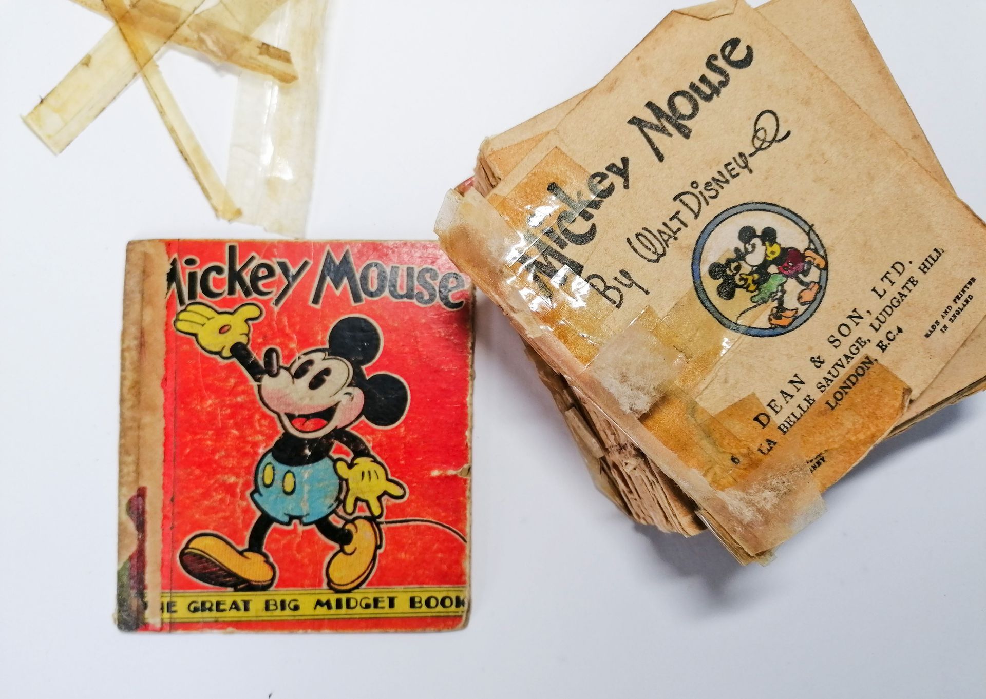The image shows a small red vintage book from the 1950's with Mickey Mouse on the cover. It is laid on a white background. Next to it is some old pieces of yellow sellotape and a seperate pile of stacked pages which are old and torn and are covered in old brittle sellotape.