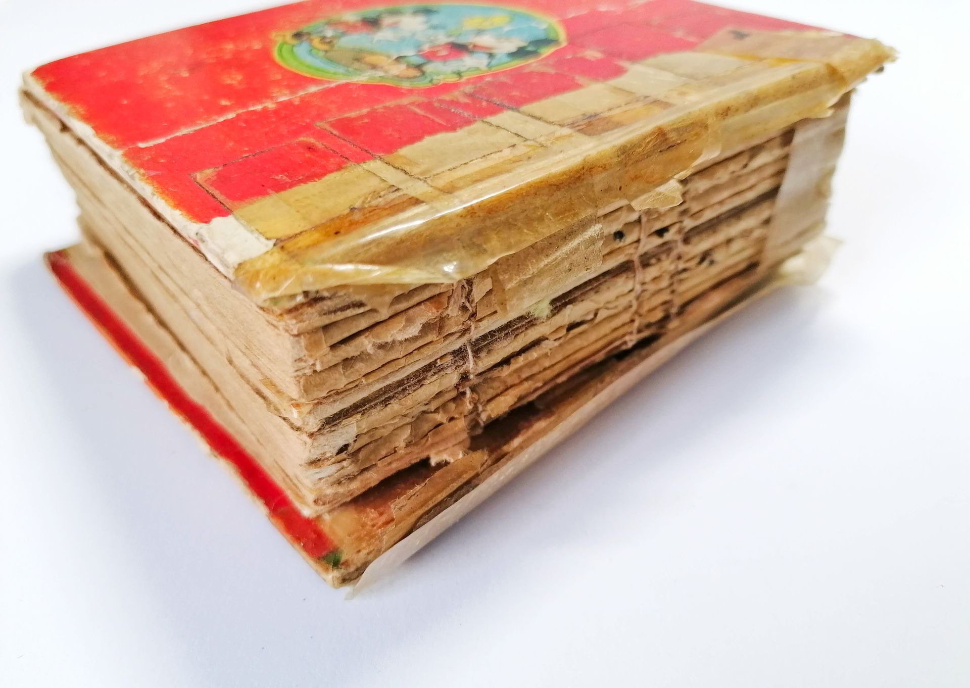 An image of a small damaged book needing repair. The spine of the book is shown in detail where the pages are rotten and damaged and pieces of old yellow dried sellotape are seen holding the covers onto the book block. The back cover of the book is red and there is an image of Mickey and Minnie mouse.