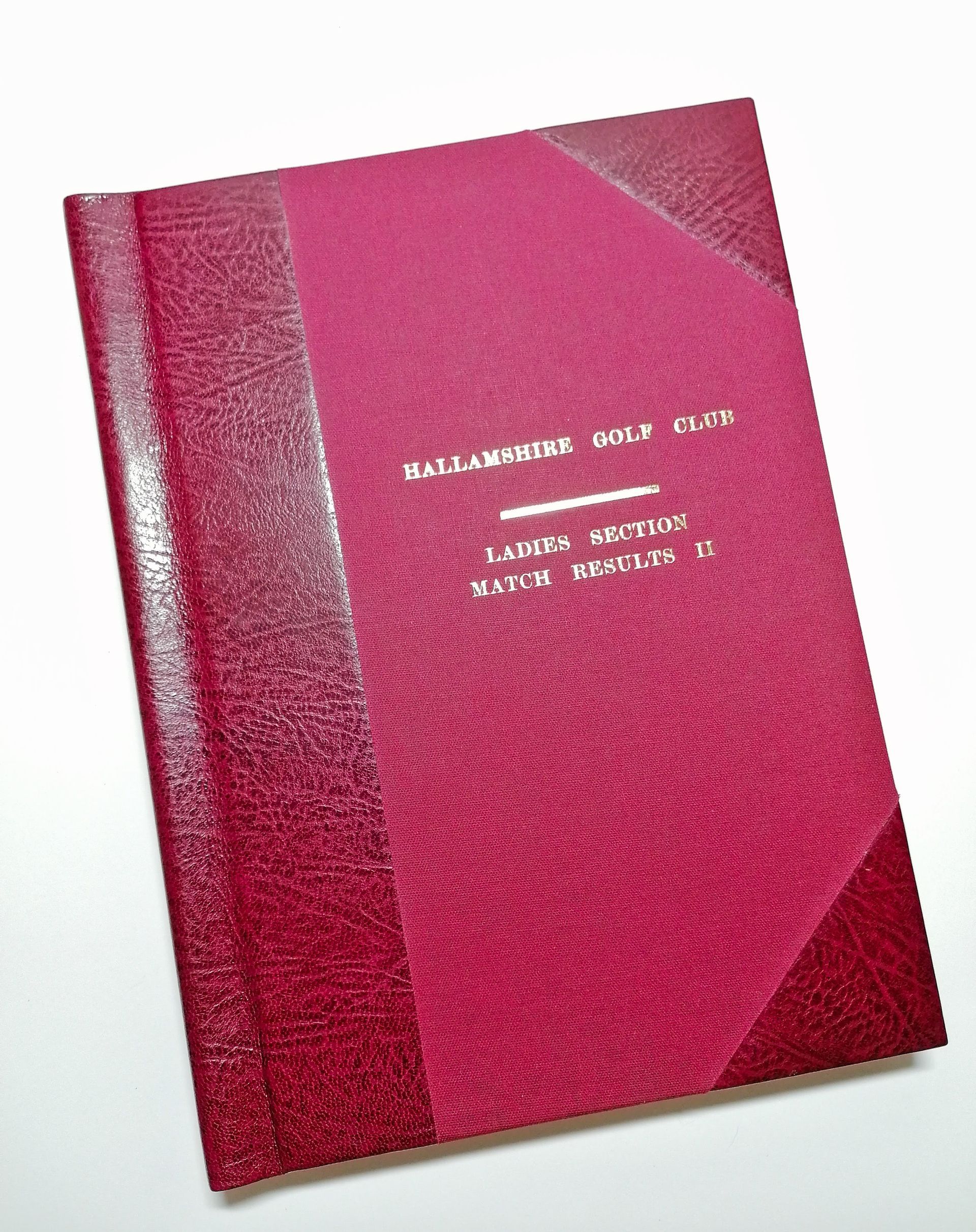 The image shows a large A4 sized portrait red leather and cloth golf book. The book has a leather spine and leather corners. Two on the front and two on the back. The front and back sides of the covers are covered in red book cloth. There is gold embossed lettering on the cover which reads Hallamshire Golf club, underlined with a gold line. The second line reads Ladies Section and the third line reads Match Results two, in roman numerals. The book is laid on a white background.