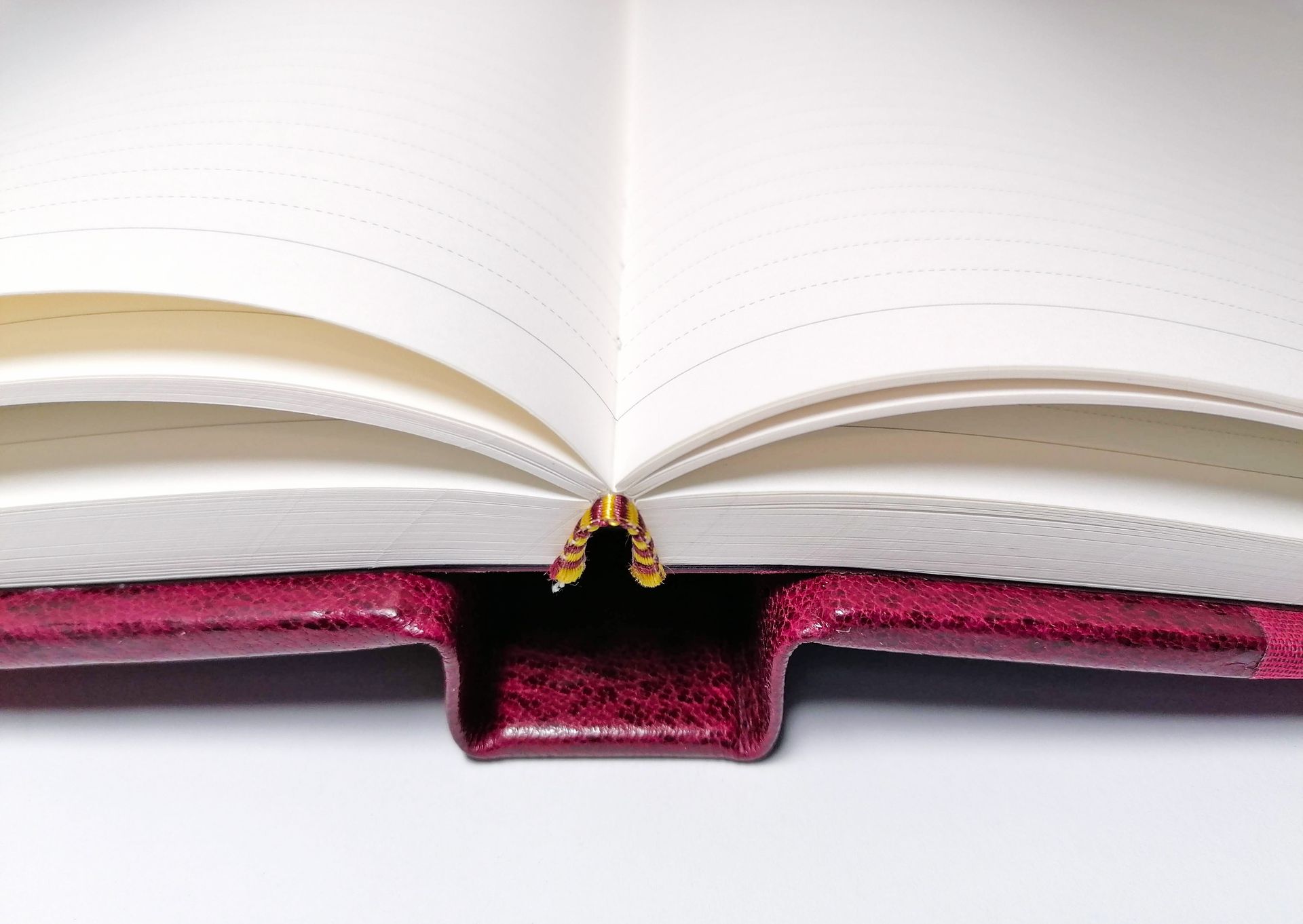 The image shows a red leather book opened flat on a white background. The pages are light cream coloured and have faint grey lines for writing onto. The book is parted in the middle so the pages lays flat and there is a coloured fabric headband on the top of the pages which is red and yellow.