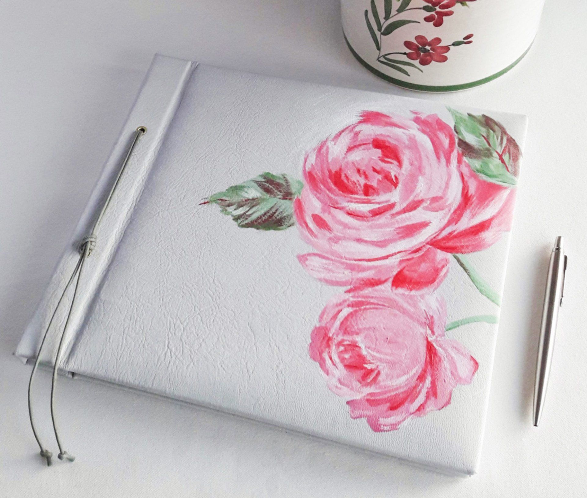 The image shows a light grey 100% leather landscape wedding guest book with roses painted by hand on to the front cover. The roses were painted by Holly of bespoke bindings and are in Light pink, dark pink and the leaves are in various greens.