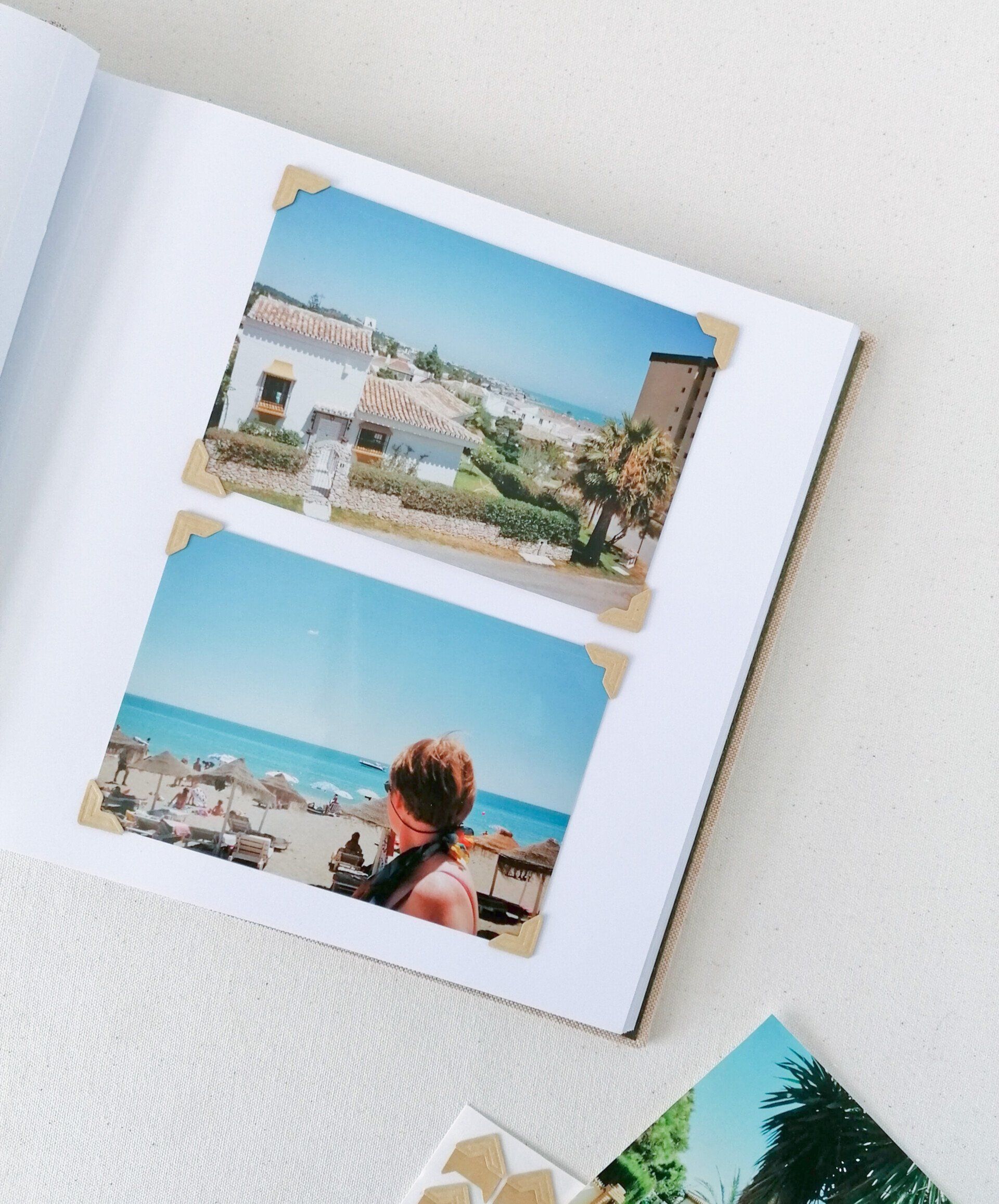 An image of a peach linen landscape photo album. The album is laid open with white pages. One page has 2 landscape holiday photos. the one on the top of the page is of a villa in Spain and the surrounding landscape below against a blue sky. The photo underneath is of a blue sky against a sandy beach and a lady has her back turned towards the camera and is looking down the beach at the straw umbrellas and sun loungers. The album is laid on a white background. The photos are attached to the white page at the corners, with 4 brown craft card photo corners per photo.