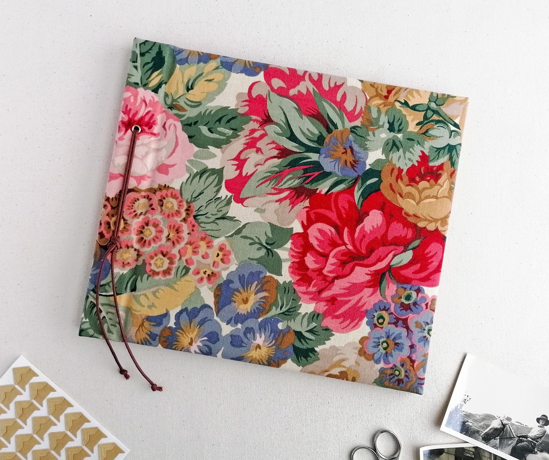 The image shows a vintage Sanderson floral fabric covered album. The decorative fabric is floral with red, pink, green, blue and mustard flowers on a cream background. The cord tying the album together is red.