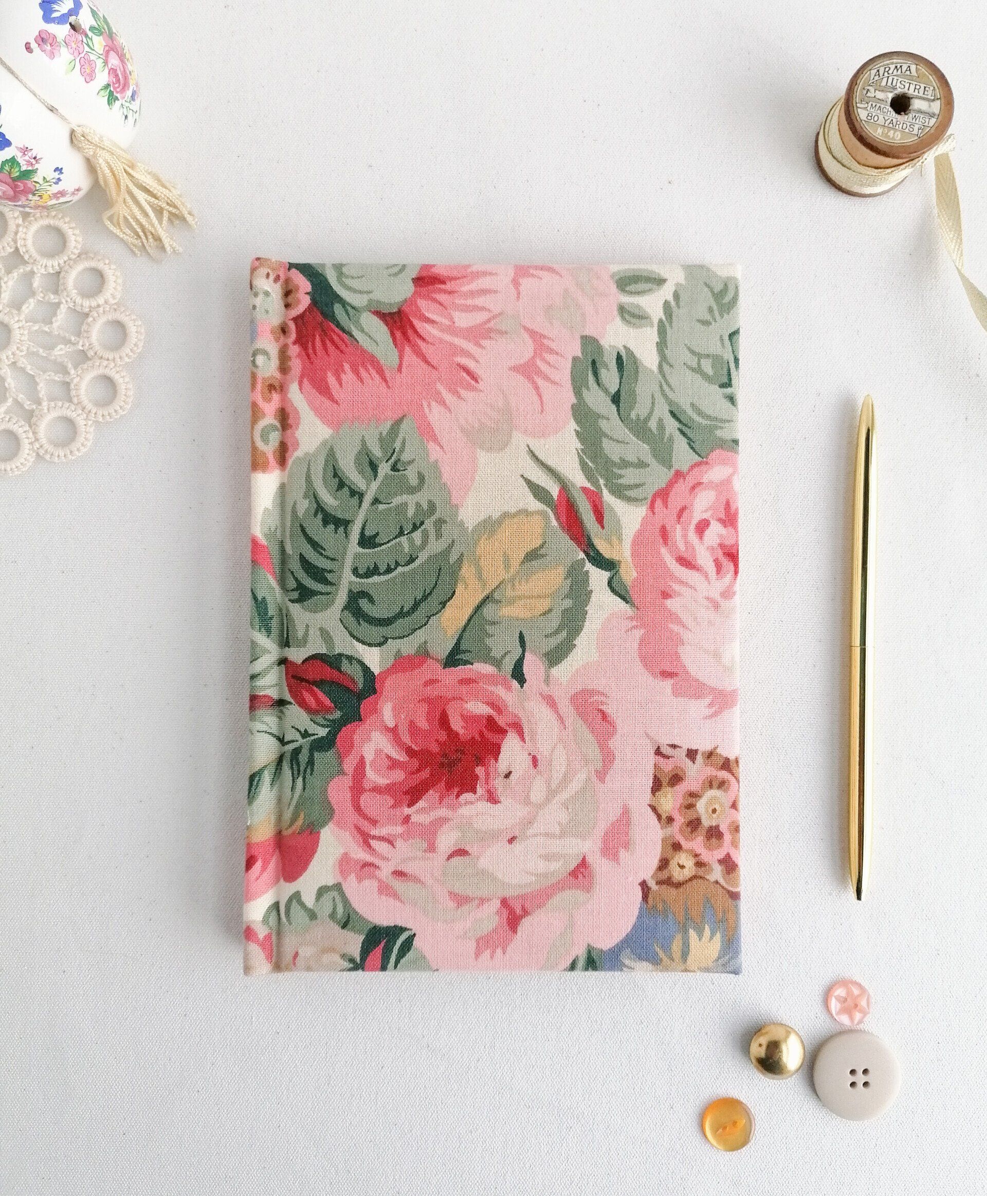 An image of a vintage Sanderson fabric A5 address book with padded covers. The floral pattern on the covers is in red light pink dark pink green blue and mustard. It is laid on a plain white background. There is a gold pen laid next to the book.