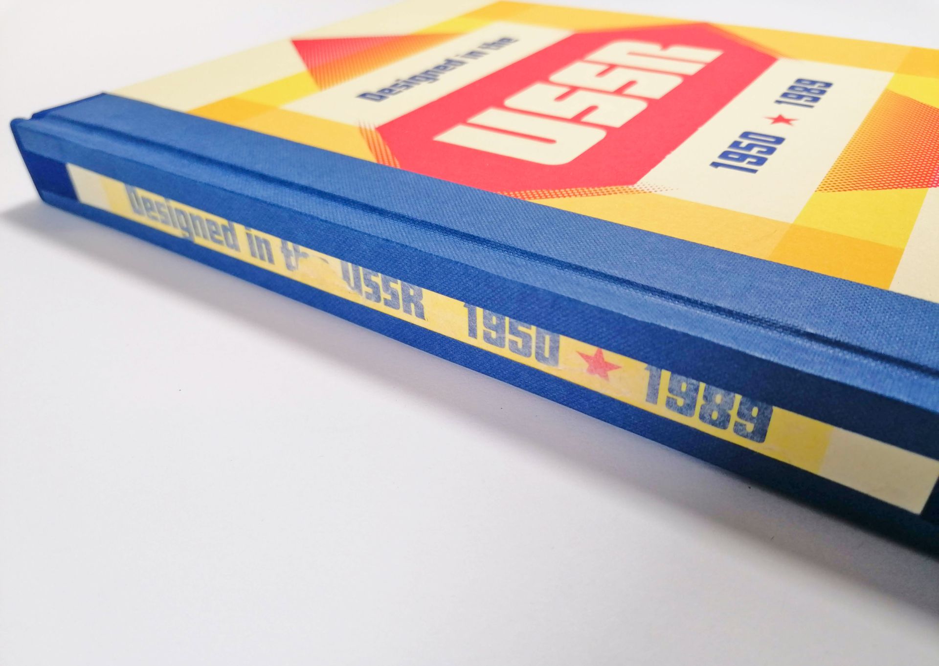 An image of the repaired blue and yellow hard back book called Designed in the USSR 1950 TO 1989. The paper covered spine board is now in place and has been reattached to the covers of the book using a dark blue book cloth and blue paper hinge strip on the front and back of the book.