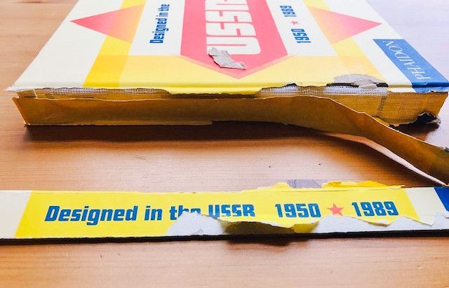 An image of a damaged blue and yellow hard back book called Designed in the USSR 1950 TO 1989. The paper covered spine board from the book is torn and seperated from the book. It is missing some of the text on the spine.