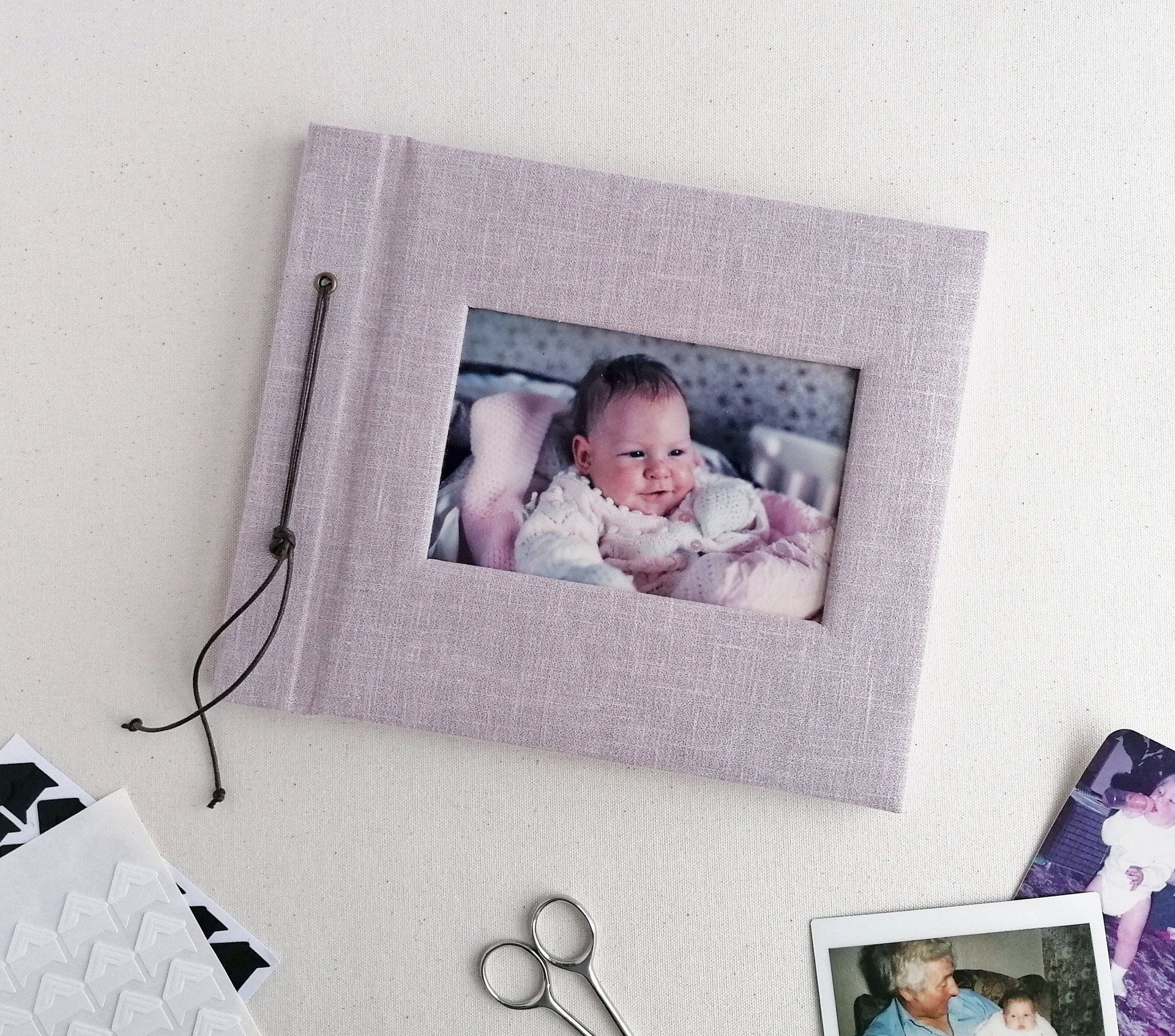 The image shows a small landscape baby photo album with a lilac cotton cover which is padded. A cut out window has a photo of a baby on the front cover.