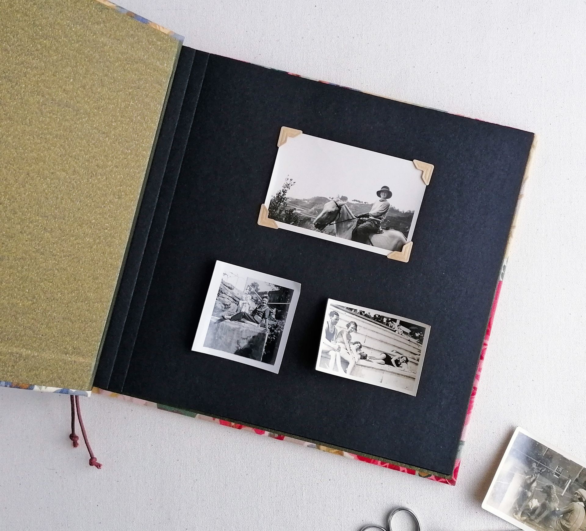 The image shows the Sanderson fabric floral album opened to the first page. The pages are black and there are 3 photos laid loosely onto the first page. The photos are vintage and in black and white showing swimmers and a lady on horseback.