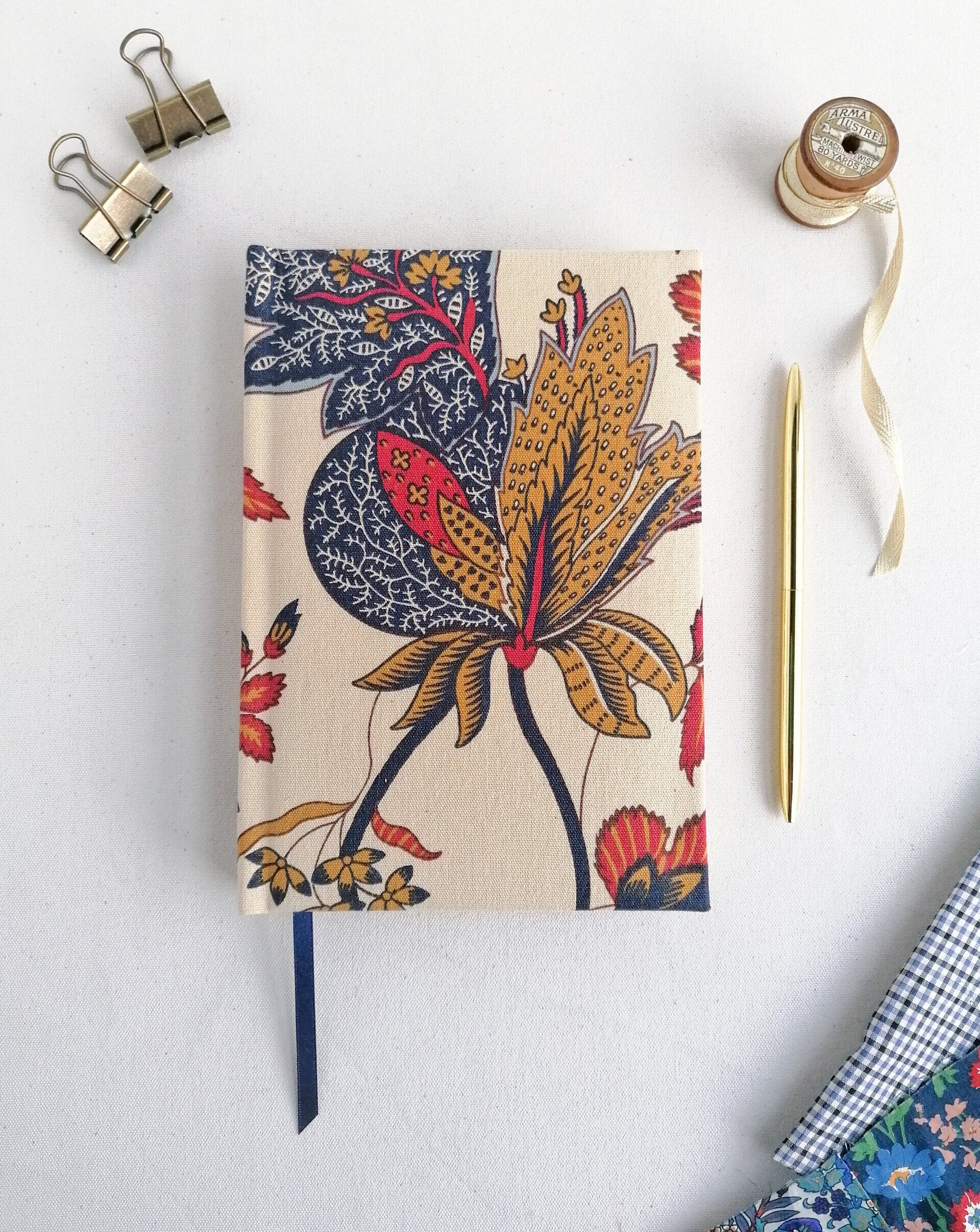 An image of a vintage Laura Ashley floral fabric on an A5 notebook with contrasting dark blue ribbon page marker. It is laid on a plain white background. There is a gold pen laid next to the book.