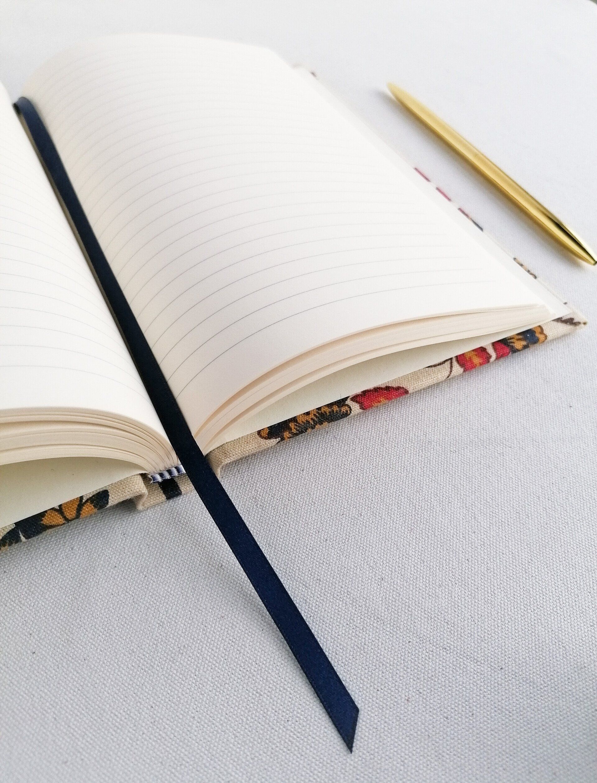 An image of a vintage Laura Ashley floral fabric on an A5 notebook with contrasting dark blue ribbon page marker. The book is laid open with grey feint lined white paper pages. It is laid on a plain white background. There is a gold pen laid next to the book.