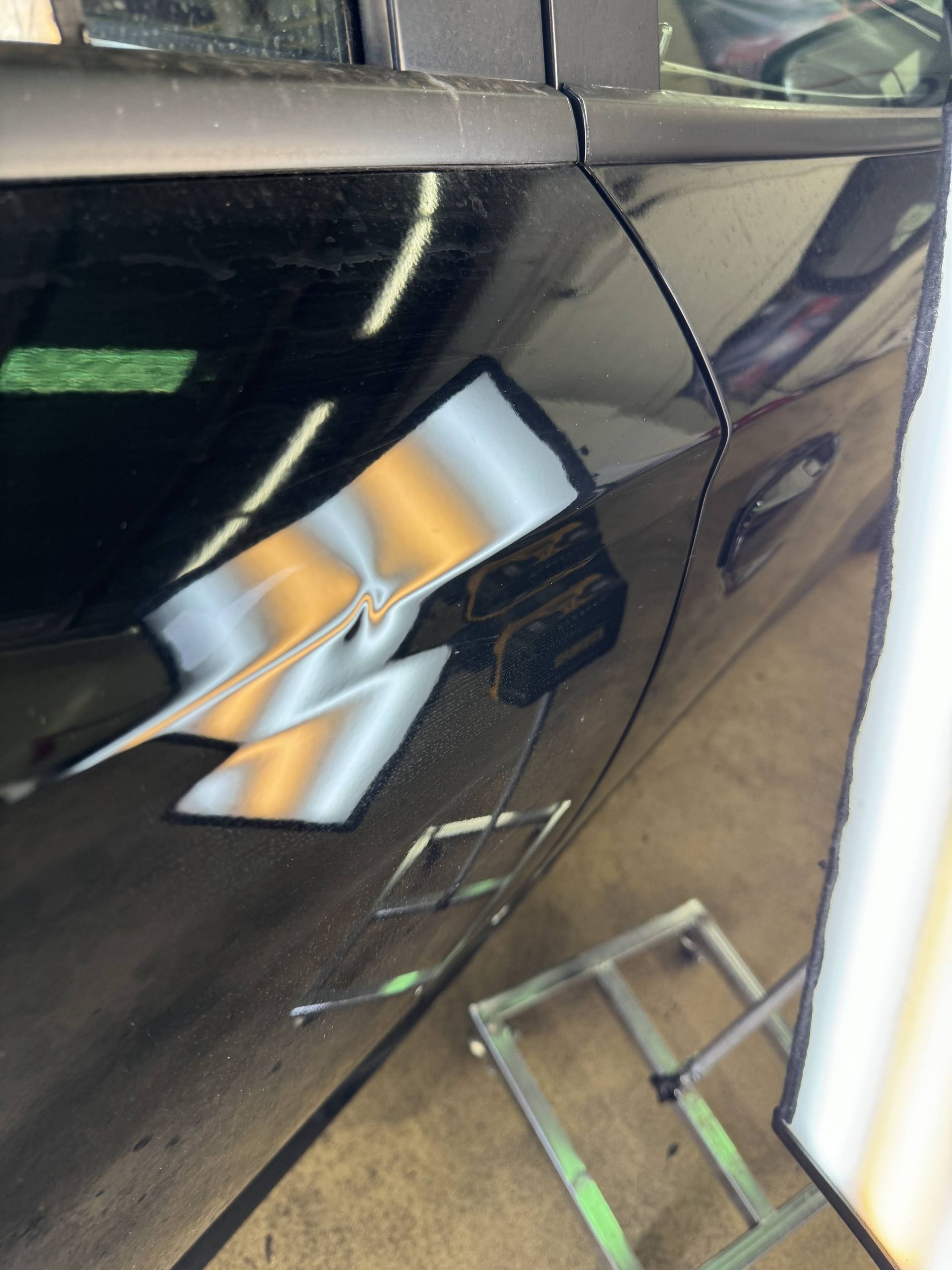 Close-up of a glossy black car door with bright light reflections and a ladder reflected below.