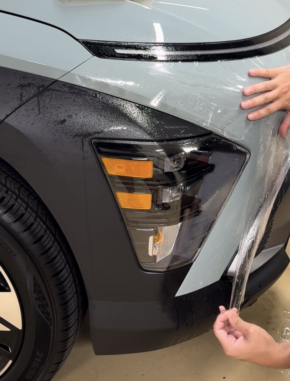 Hands peel clear protective film from a car’s front fender and headlight area.