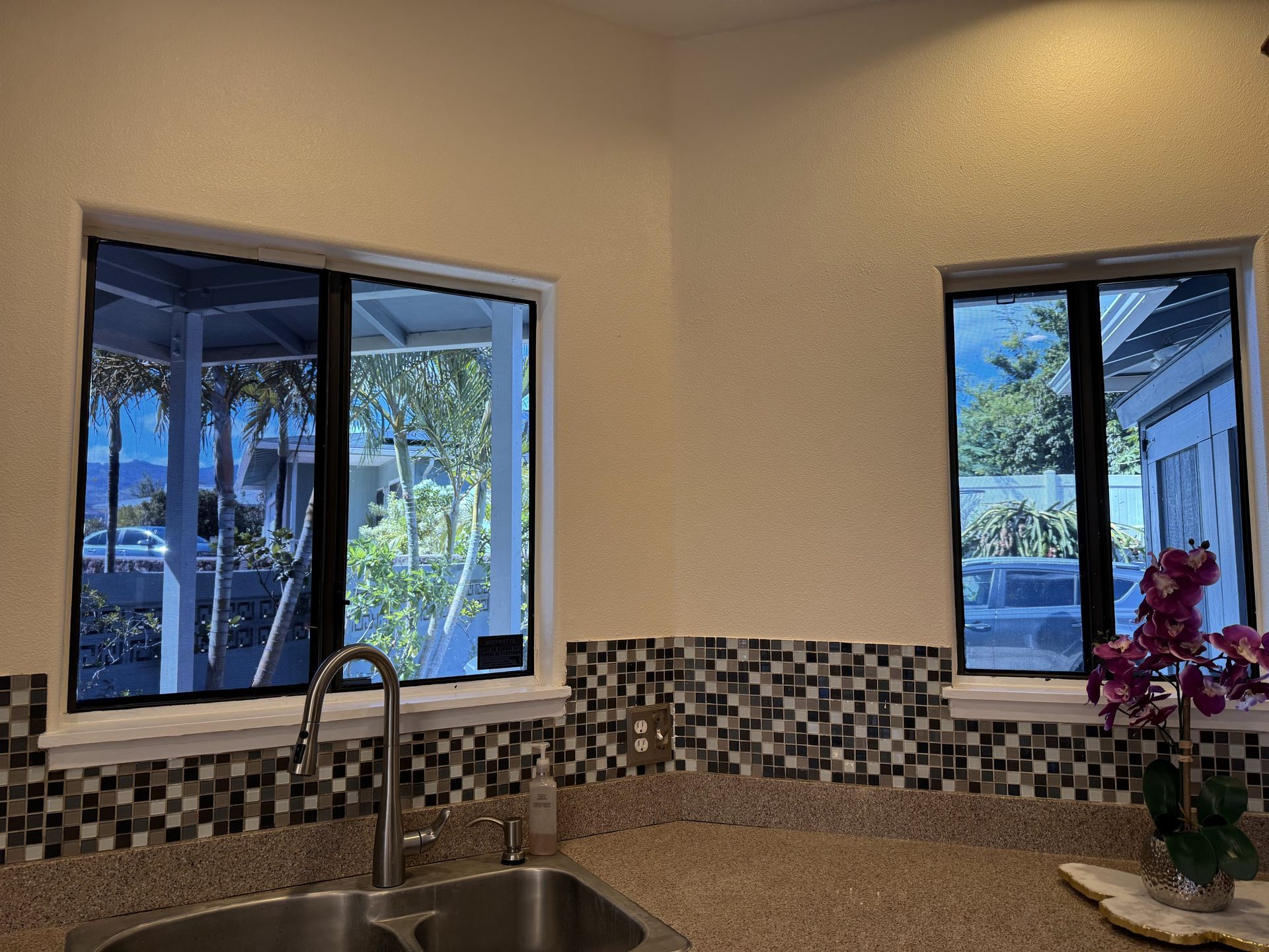 Kitchen sink beneath two windows overlooking a backyard, with mosaic tile backsplash and a flower vase on the counter.