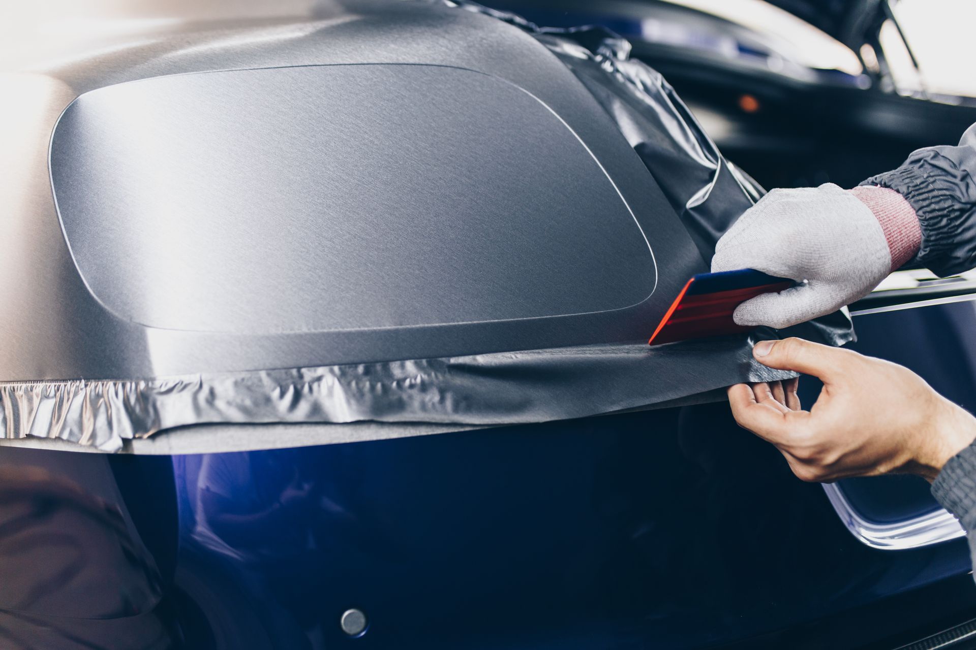 Hands applying silver vinyl wrap to a car bumper in a workshop