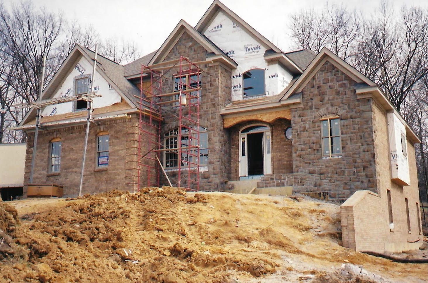 House under construction with stone exterior, scaffolding, and dirt in front.