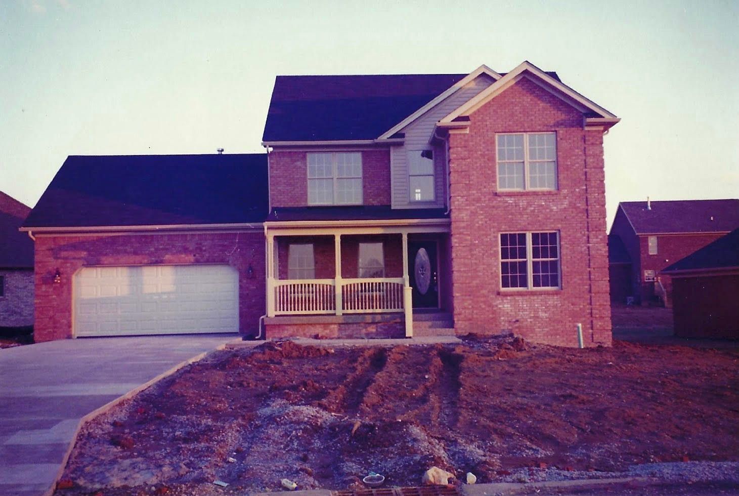 Red brick two-story house with black roof, attached garage, and unfinished yard; daytime.
