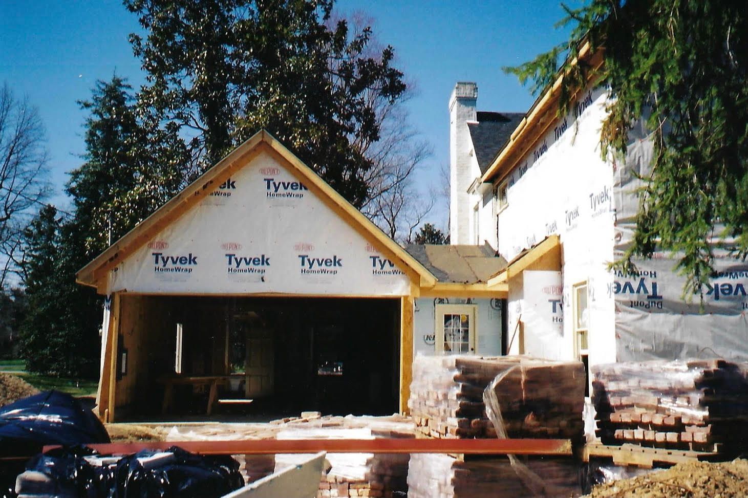 Garage under construction, attached to a house. Framing, Tyvek wrap, and stacked lumber are visible.