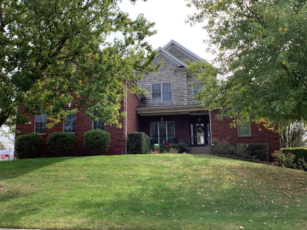 Two-story brick house with stone upper section, porch, and green lawn; framed by trees on a sunny day.