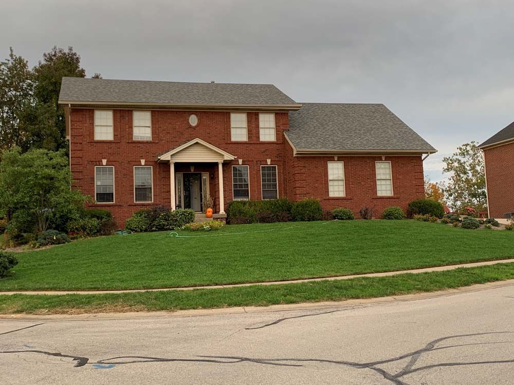 Brick two-story house with gray roof, green lawn, and cloudy sky.