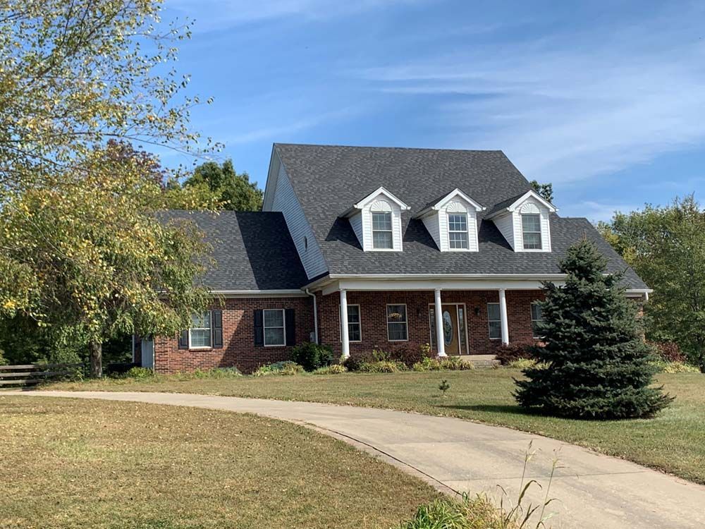 Brick house with dark roof and three dormer windows, blue sky. Long driveway.