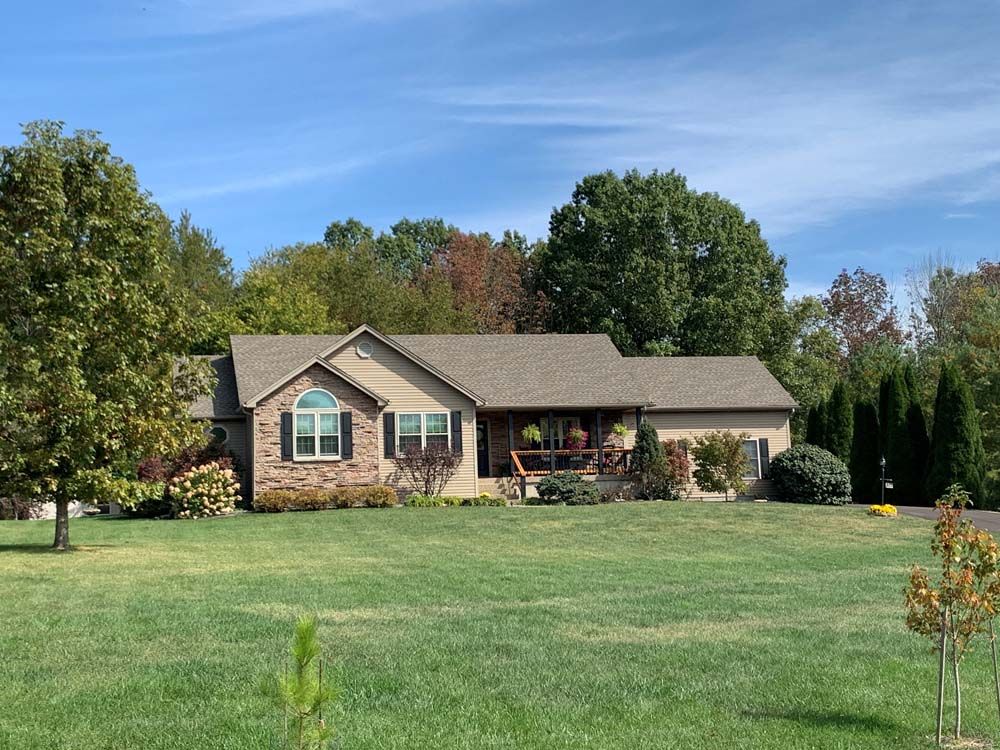 Ranch-style house with tan siding, brick facade, and a covered porch on a green lawn with trees and a blue sky.