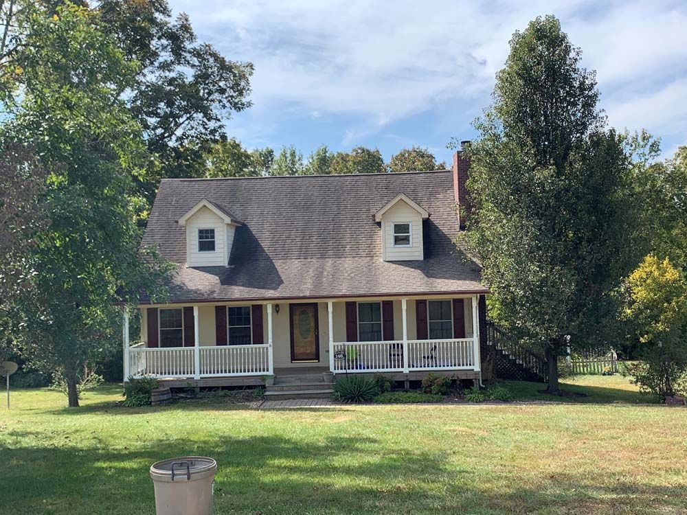 Small house with a porch, dormers, and red shutters, nestled amongst trees and a grassy lawn.