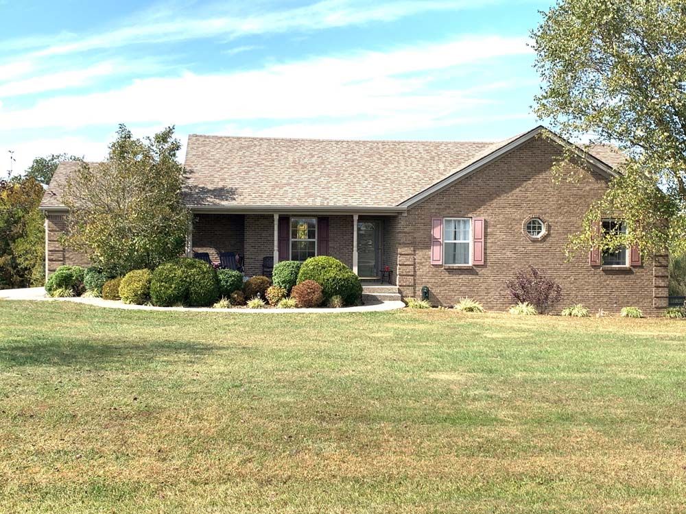 Brick house with brown roof, front porch, and lawn. Blue sky with trees on either side.