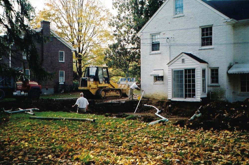 Construction site: Workers near a house, bulldozer, and a dug-out area with pipes. Autumn leaves.