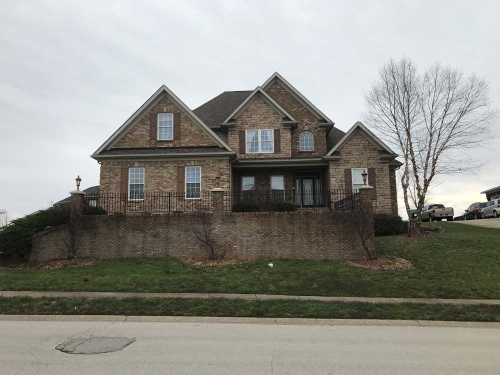 Two-story brick house with multiple gables on a grassy hill; a bare tree is to the right.