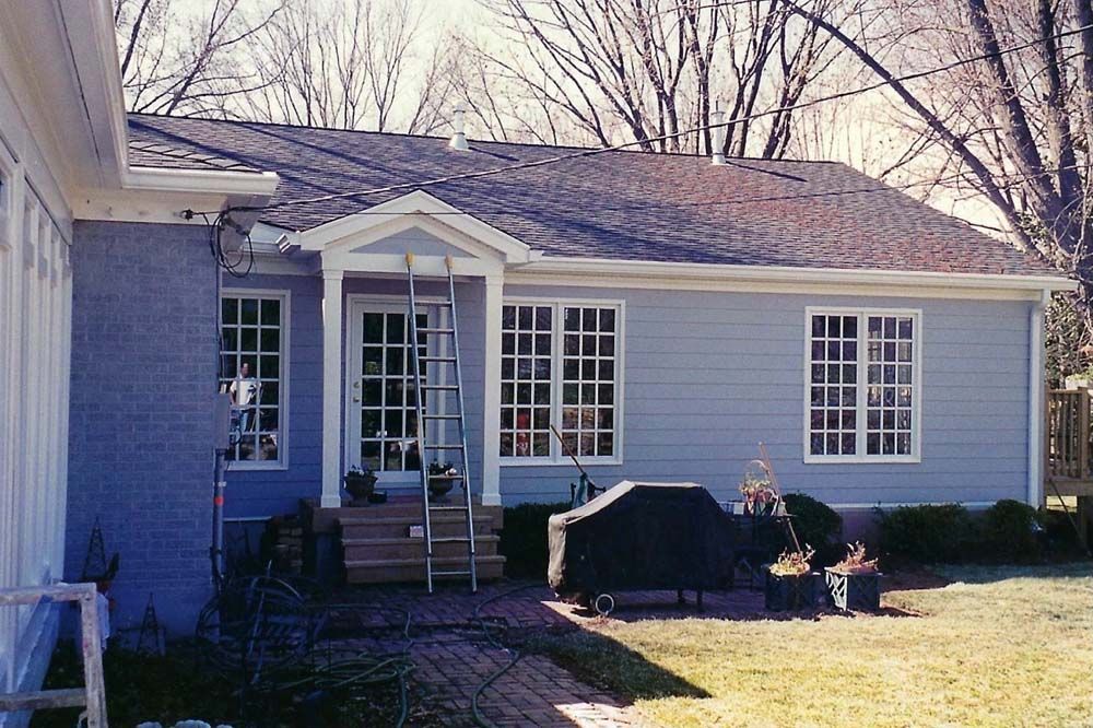 Blue house with white trim, windows, and a doorway. Backyard patio, grill, and shrubs in view.