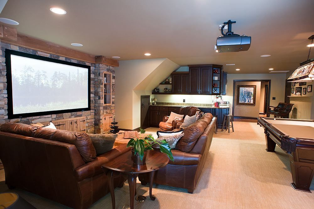 Cozy home theater with a projection screen, brown leather couches, and a pool table. Warm lighting and stone accent wall.