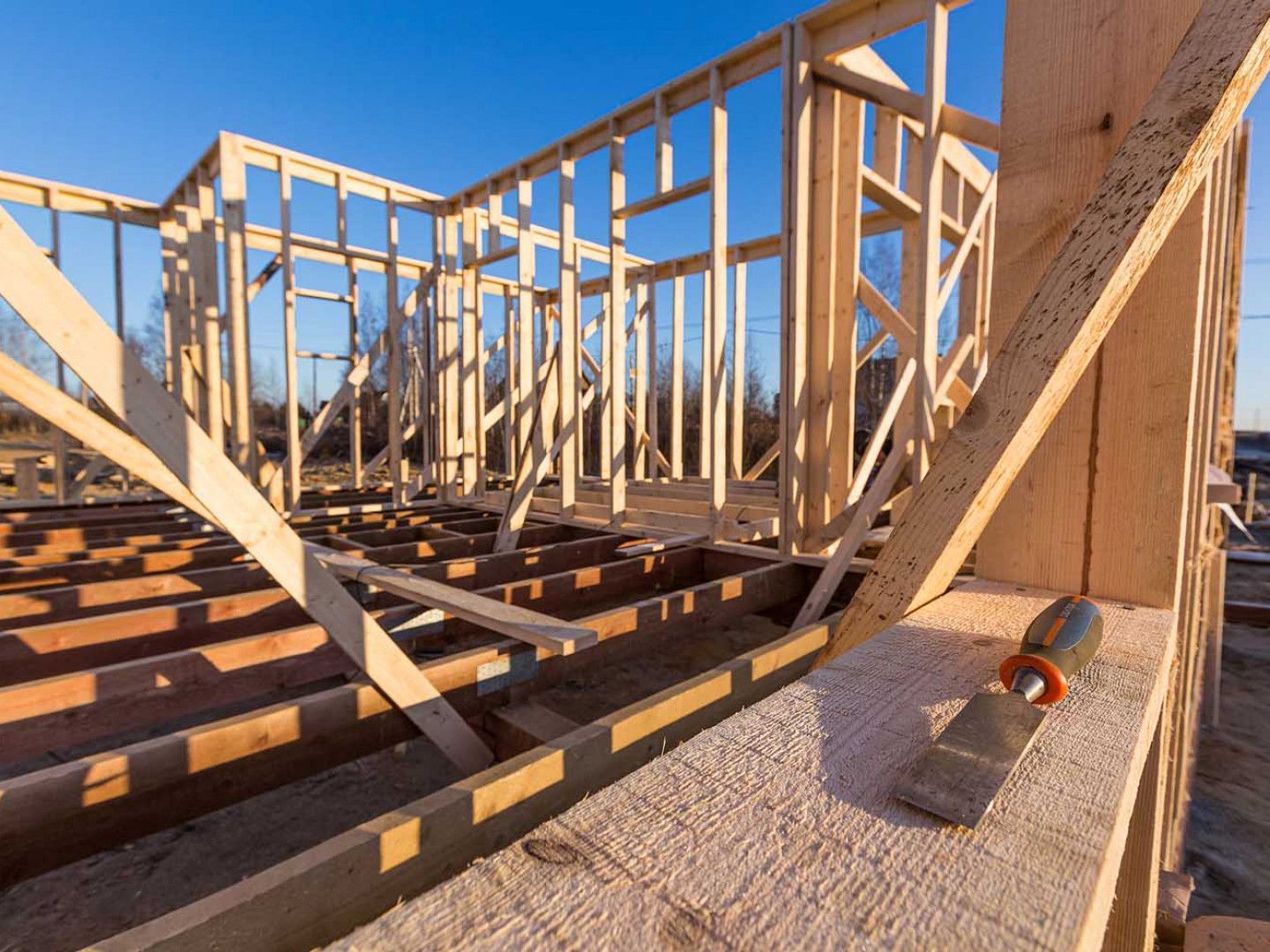 Wood frame of a house under construction with a clear blue sky in the background. A tool rests on a beam.