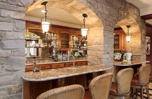 Stone-walled home bar with arched openings, granite countertop, bar stools, and liquor display.