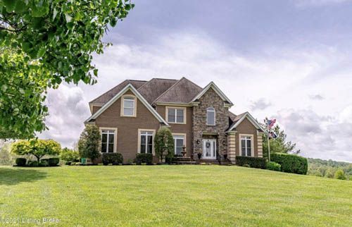 Large brown brick house with stone accents, on a grassy hill under a cloudy sky.